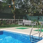 A small backyard pool with clear blue water, surrounded by a tiled deck and a grassy area. In the background, there is a white outdoor bench and chairs set, trees with colorful hanging lights, and a stone wall with green fencing for privacy.