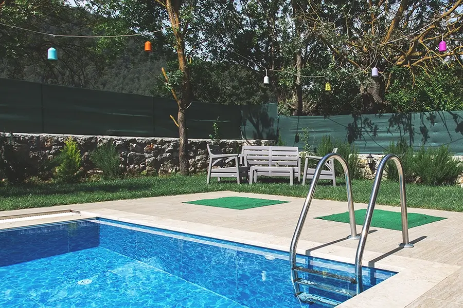 A small backyard pool with clear blue water, surrounded by a tiled deck and a grassy area. In the background, there is a white outdoor bench and chairs set, trees with colorful hanging lights, and a stone wall with green fencing for privacy.