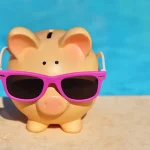 Piggy bank with sun glasses on the concrete next to pool water.
