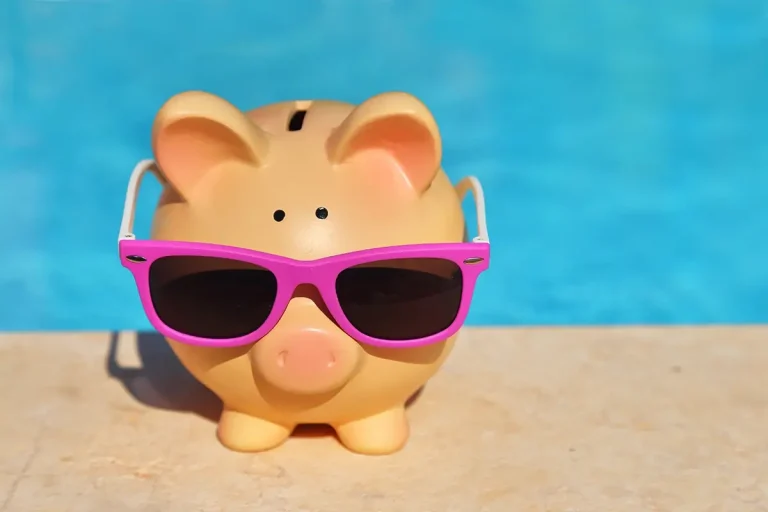 Piggy bank with sun glasses on the concrete next to pool water.
