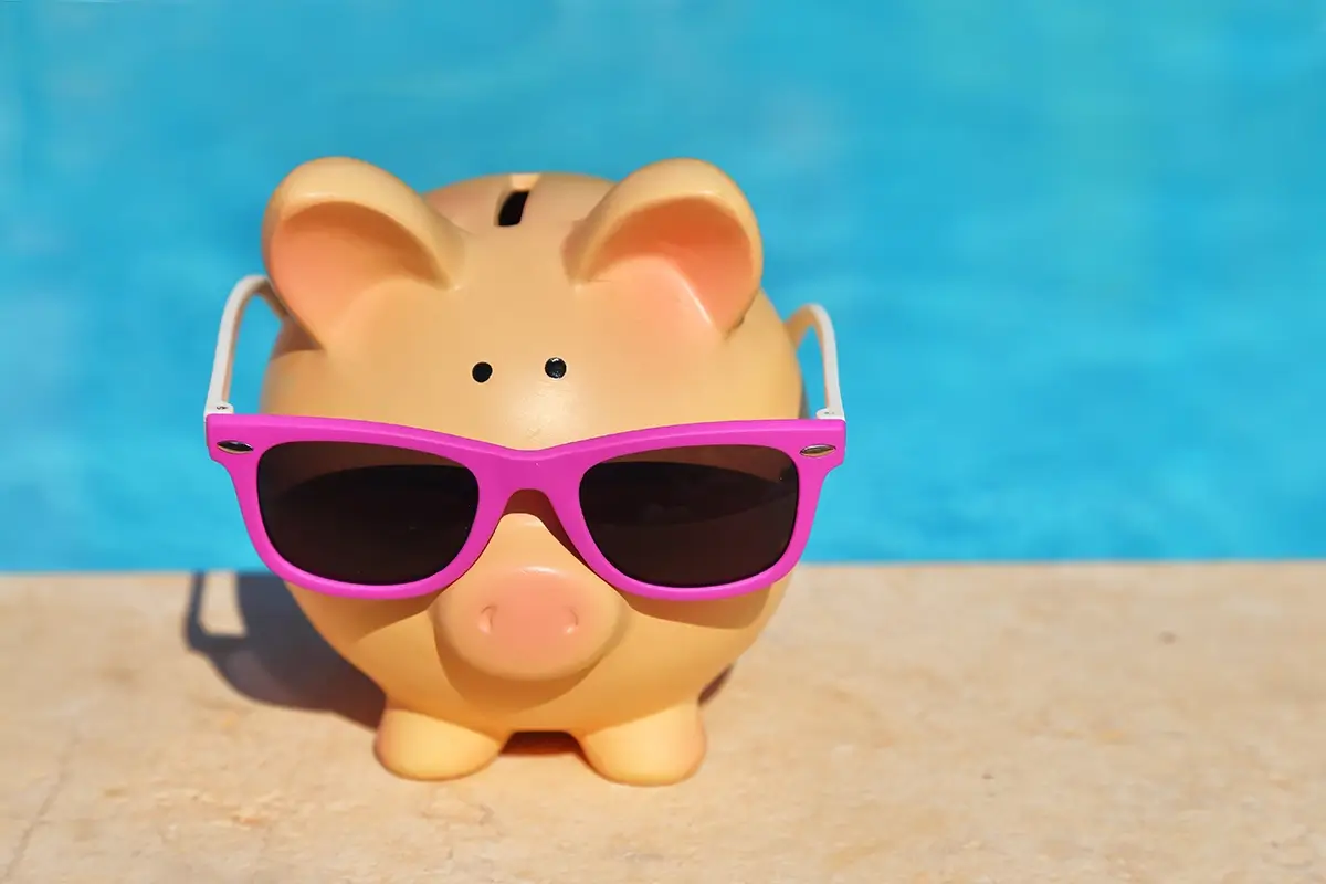 Piggy bank with sun glasses on the concrete next to pool water.